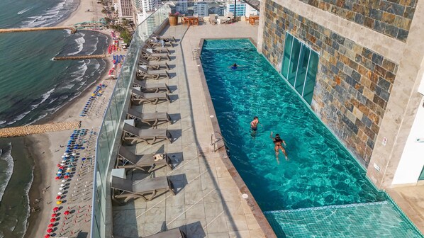 Una piscina en la azotea con agua cristalina, junto a una playa con sombrillas y sillones de descanso.