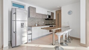 A modern kitchen with a stainless steel refrigerator, white cabinets, granite countertops, a built-in oven, and a bar area with white stools. - Ocean Bliss 2BR - Luxury Apartment | Bocagrande, Cartagena (Cartagena de Indias)