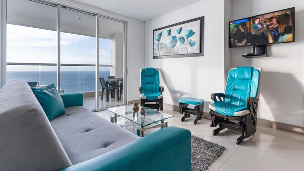 A modern living room with a sofa, teal chairs, a glass coffee table, and a TV on the wall.
