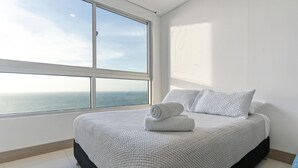 A bedroom with a large window offering a view of the sea, a neatly made bed with white bedding, and a clean, minimalist design.