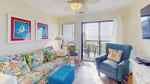 Un salon avec un canapé aux couleurs vives, un fauteuil bleu, un ventilateur de plafond et une vue sur un espace extérieur, accessible par une porte vitrée.