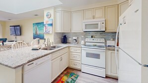 Une cuisine avec des meubles blancs, un four à micro-ondes, un four et un lave-vaisselle. Il y a un tableau au mur et un tapis coloré sur le sol.