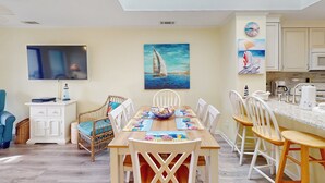 Un coin repas avec une table en bois, des chaises blanches et un grand tableau représentant un voilier accroché au mur.