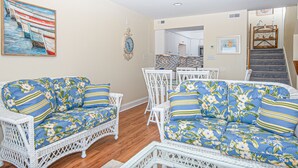 A living room with wicker furniture, a dining table, and a kitchen in the background. - Multi-Level, Oceanfront Condo in Pine Knoll Shores w/Free WiFi, Shared Pool (Pine Knoll Shores)