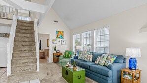 A spacious living room with a staircase, a blue-striped sofa, a green chest of drawers, and a window with blinds.
