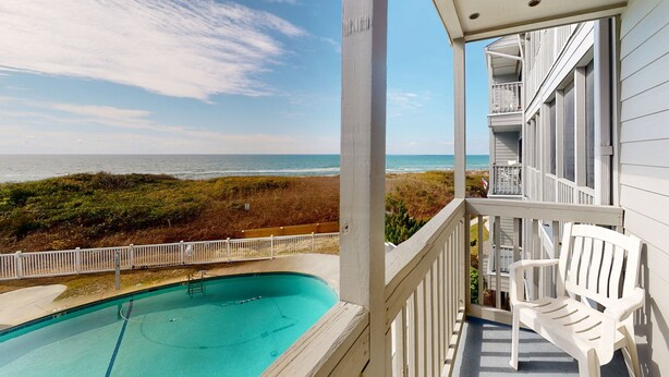 Ein Pool mit Blick auf Strand und Meer, ein weißer Stuhl und ein Balkon mit Geländer.