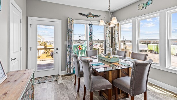 A dining area with a wooden table, grey upholstered chairs, and a chandelier. - Ocean Crystal A: 2nd-row duplex, ocean views, walk to beach/pier (Atlantic Beach)