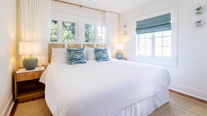Un dormitorio ordenado con una cama grande, dos mesitas de noche, un ventilador de techo y una ventana con vistas a la vegetación.