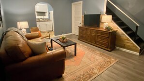 Una sala de estar con un sofá, una mesa de centro y un televisor sobre un mueble de madera. Se ve una escalera a la derecha y una cocina al fondo.