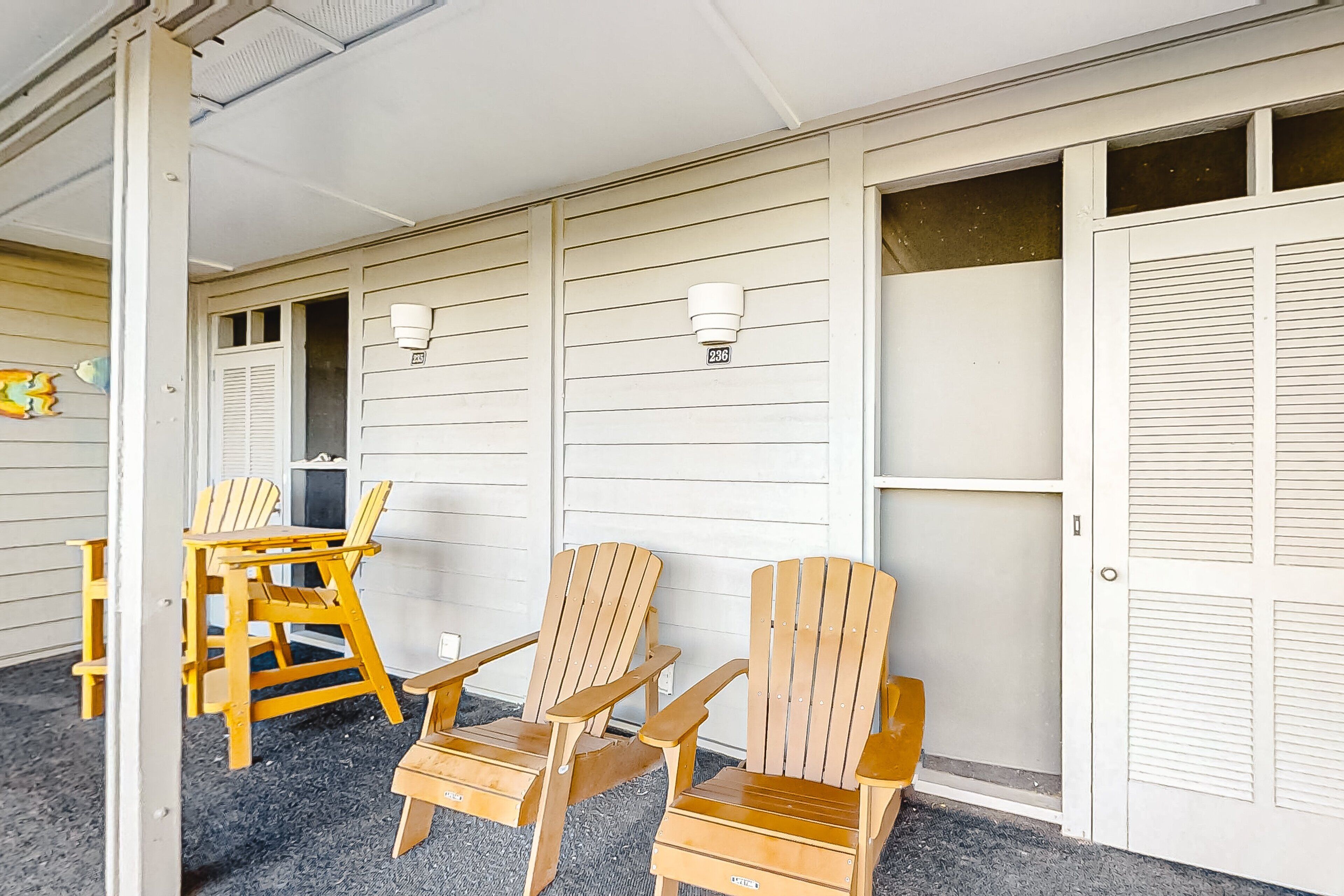 Une véranda couverte avec des chaises et une table en bois, des murs recouverts de bardage blanc et une porte avec une fenêtre.