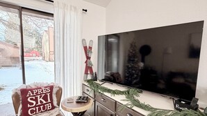 A cozy living room with a large flat-screen TV, a leather armchair with a ski club pillow, a small round table, and a Christmas tree on the mantel. - Cozy Condo -Walk to Downtown McCall & Marina! Hot Tub, Indoor Pool, Sauna &more. (McCall)