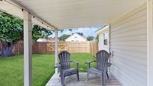 A covered porch with two Adirondack chairs facing a wooden deck and a house in the background.