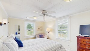 A bedroom with a bed, a ceiling fan, a dresser, a TV, and two windows with blinds.