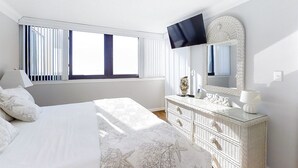Un dormitorio con cama, cómoda, televisor montado en la pared y ventana con persianas.