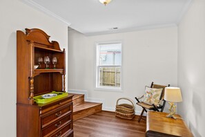 Une pièce comprenant une commode en bois, un panier en osier, une chaise avec un coussin à motifs floraux et une table avec une lampe.