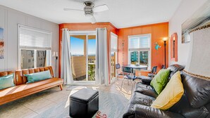 A living room with a sofa, ottoman, and a coffee table. There is a ceiling fan, a dining table with chairs, and a view through the glass doors. - Beachfront condo with indoor/outdoor pools, hot tubs, waterslide, & putt-putt (Atlantic Beach)