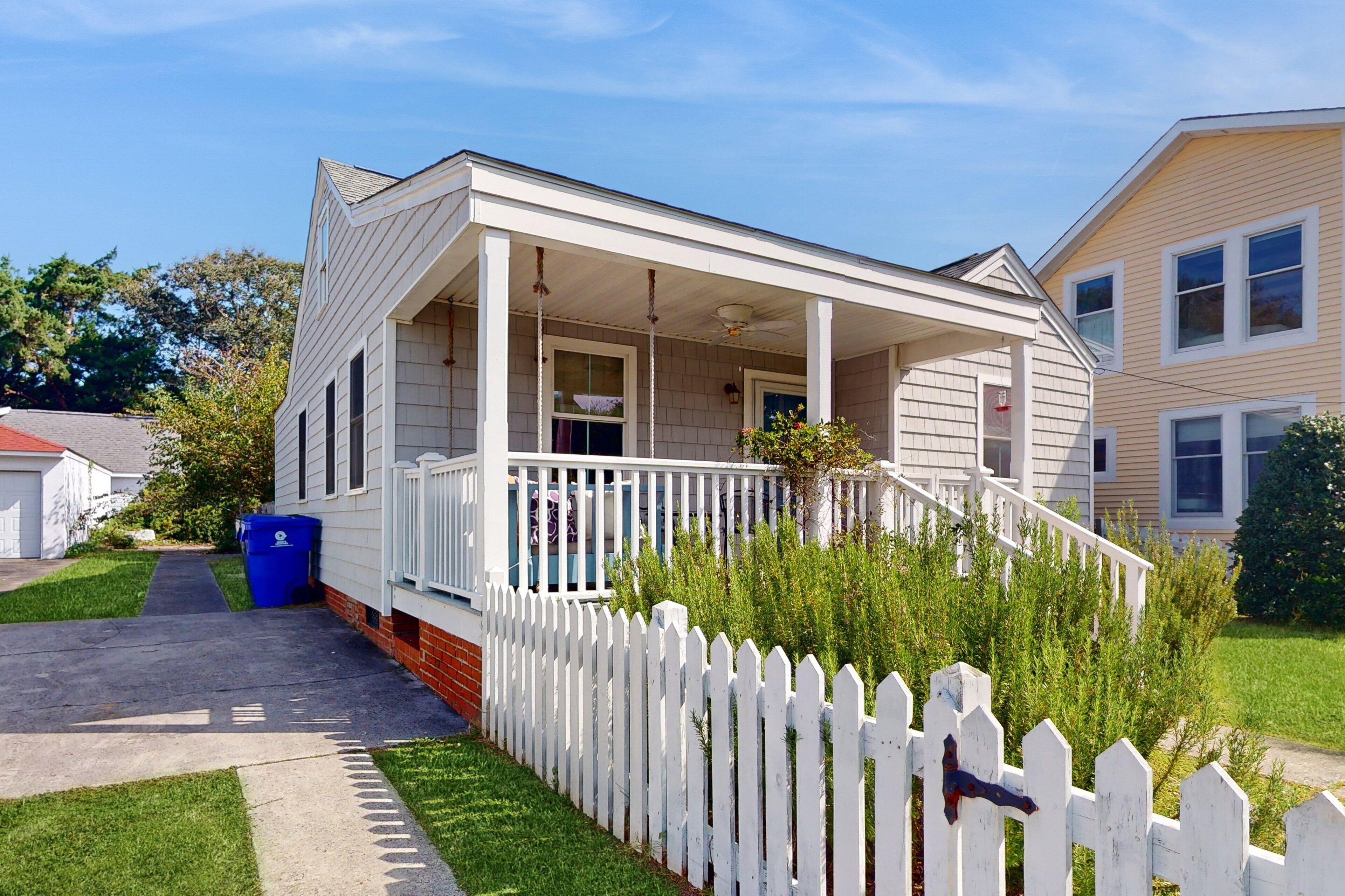 A two-story house with a front porch, a white picket fence, and a blue trash bin.
