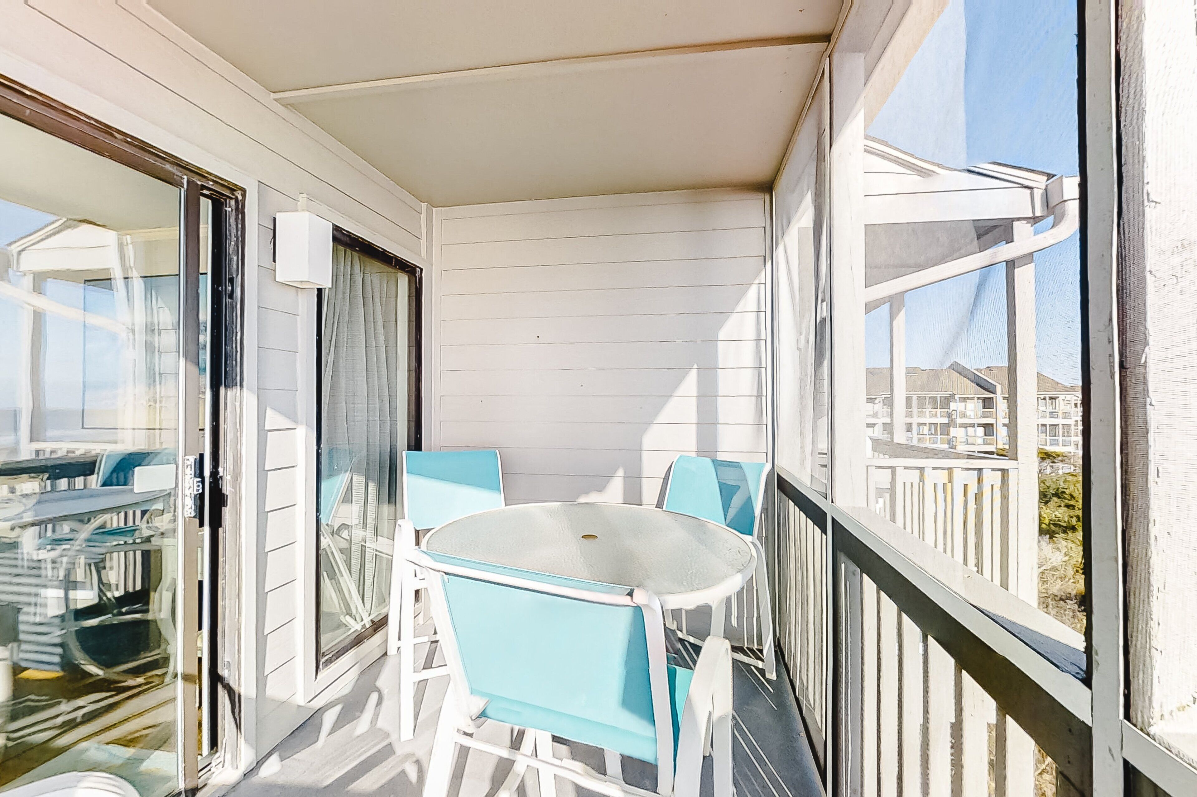 A balcony with a glass table and chairs, a sliding glass door, and a view of the beach and buildings.