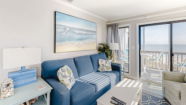 Een woonkamer met een blauwe bank, een schilderij van een strand, een tafel met een lamp, en uitzicht op de oceaan door grote ramen.