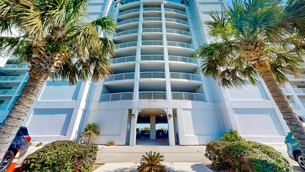 A multi-story hotel with a central entrance, surrounded by palm trees and a parking area. - Ocean view condo with shared pool, fitness room, & balcony (Indian Beach)