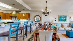 A spacious living area with a dining table, chairs, and a sofa. There is a kitchen area with wooden cabinets and a chandelier. - Ocean view condo with shared pool, fitness room, & balcony (Indian Beach)