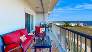 A balcony with a red sofa, a small table, and chairs, overlooking a grassy area and the sea. - Ocean view condo with shared pool, fitness room, & balcony (Indian Beach)