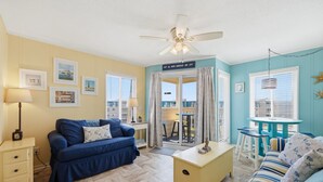 Una habitación con un sofá azul, una mesita auxiliar amarilla, un ventilador de techo y vistas a un área exterior a través de una puerta de vidrio.