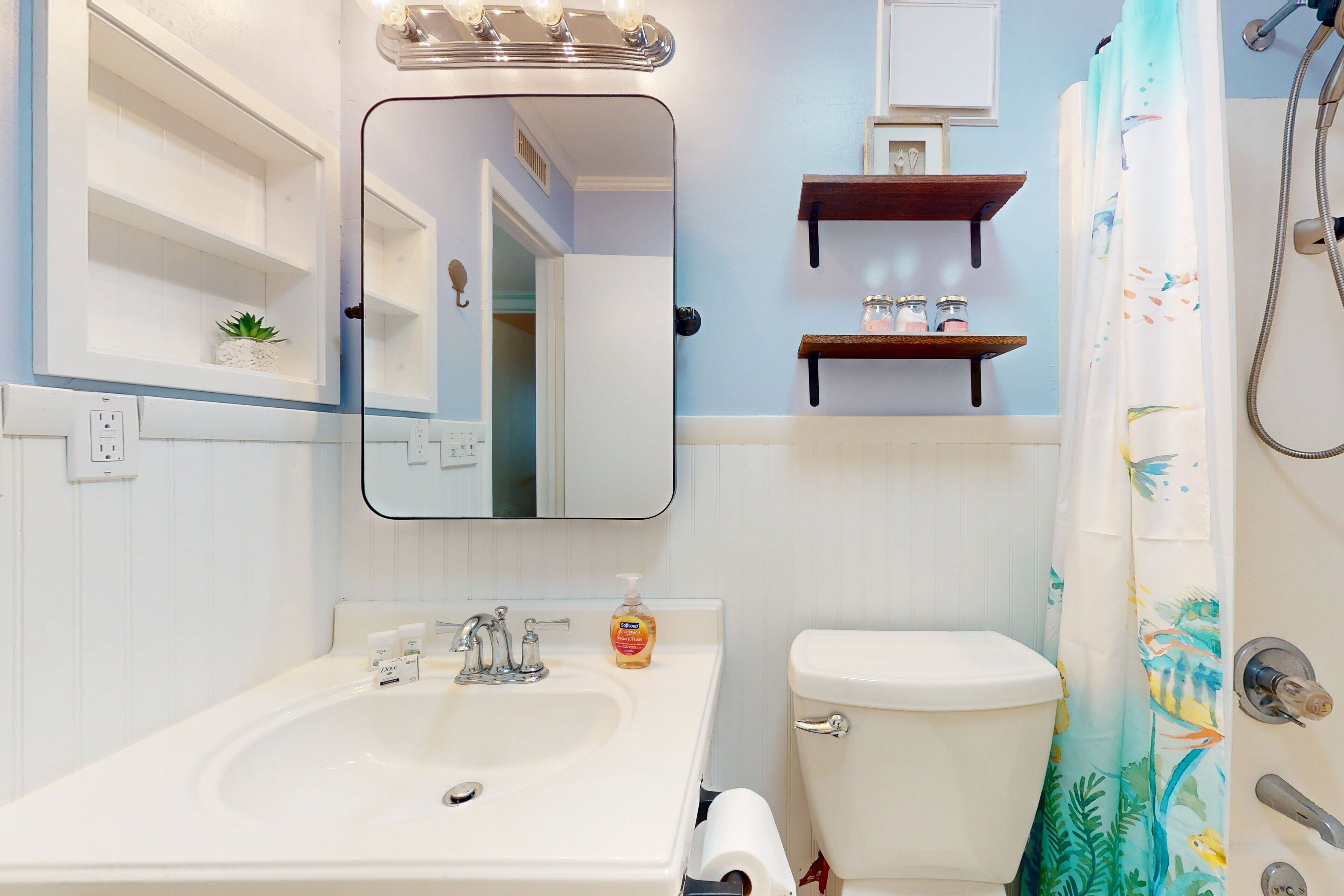 A bathroom with a sink, toilet, and shower. There are shelves above the toilet and a mirror above the sink.