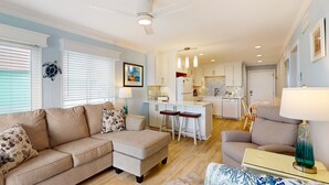 Een woonkamer met een beige bank, een klein voetenbankje en een fauteuil met een patroon. Achtergrond is een keuken met witte kasten en een eethoek met een witte tafel en stoelen.