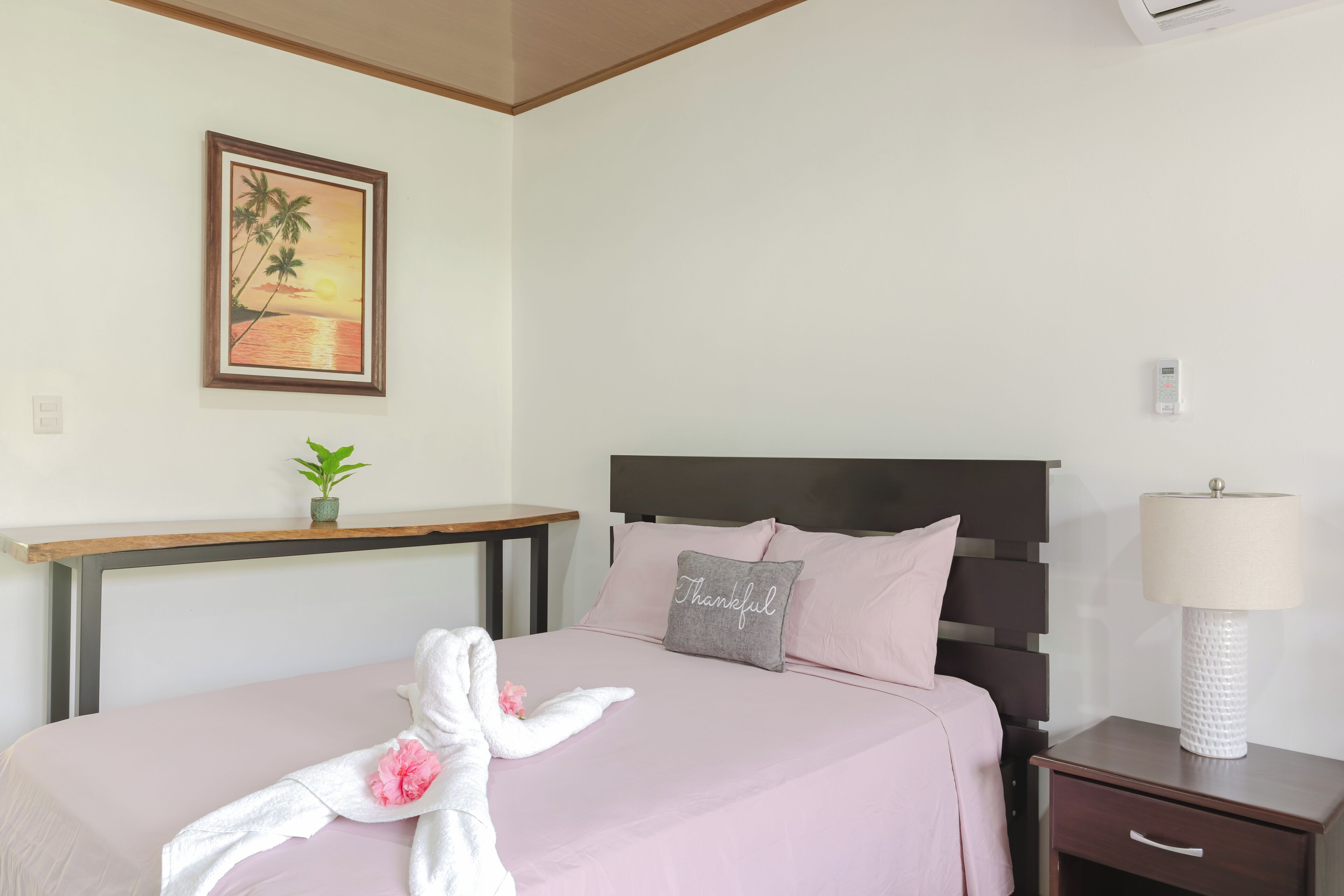Un lit bien fait, recouvert d’une couvre-lit rose, avec un oreiller sur lequel est brodé le mot « Merci », et une serviette ornée d’une fleur. Une petite plante est posée sur une étagère au-dessus du lit, un tableau représentant un coucher de soleil est accroché au mur, et une table de chevet avec une lampe se trouve à côté du lit.