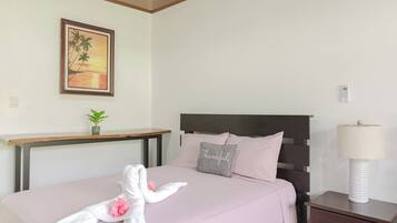 Un lit bien fait, recouvert d’une couvre-lit rose, avec un oreiller sur lequel est brodé le mot « Merci », et une serviette ornée d’une fleur. Une petite plante est posée sur une étagère au-dessus du lit, un tableau représentant un coucher de soleil est accroché au mur, et une table de chevet avec une lampe se trouve à côté du lit.