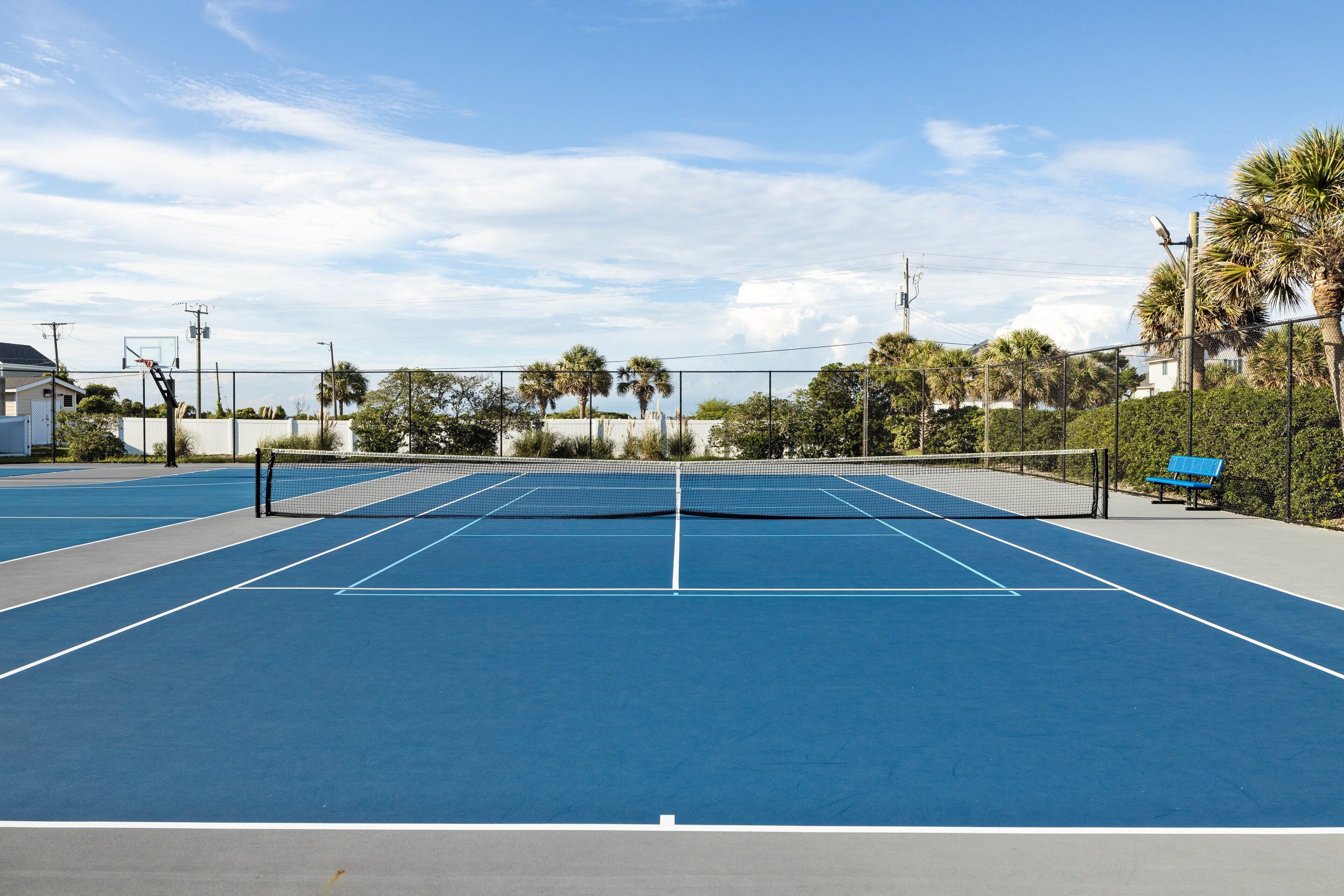 Een buitenterrein voor tennis met een net, omheind door een hek en palmbomen.