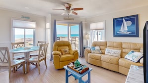 Ein Wohnzimmer mit Blick auf den Strand, einem Deckenventilator, einem Esstisch mit Stühlen und einer Couch mit Zierkissen.
