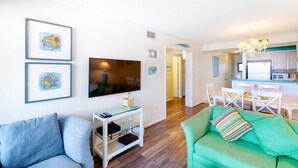 Una sala de estar con televisor, obras de arte enmarcadas, un sofá verde y una zona de comedor con sillas y mesa.