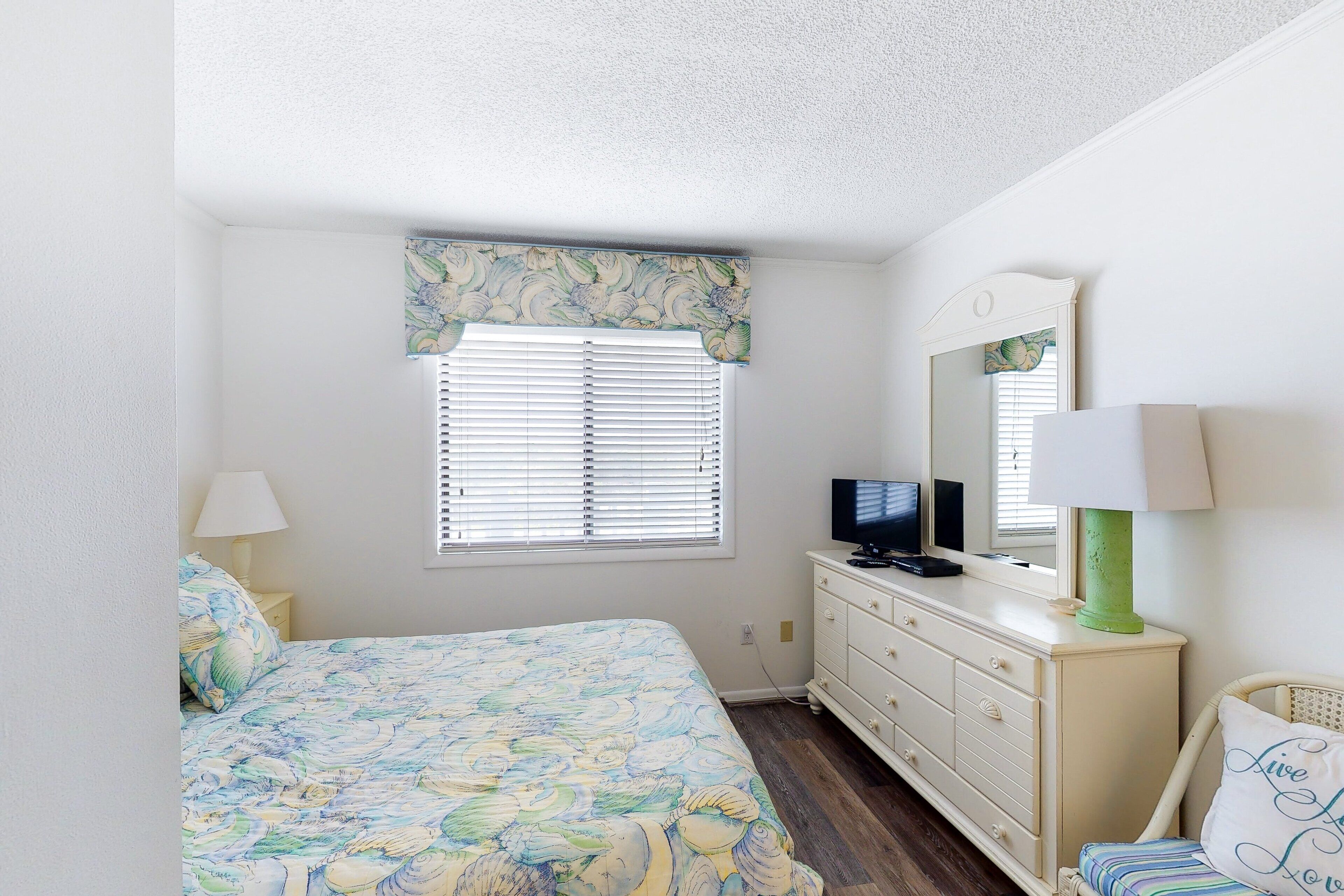 A bedroom with a bed, dresser, television, and a window with blinds.