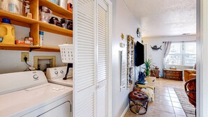 Un espace buanderie comprenant un lave-linge et un sèche-linge, des étagères avec des ustensiles de cuisine, un miroir et une fenêtre avec des rideaux.
