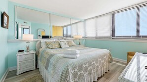A bedroom with a large mirror, a bed with a patterned bedspread, a bedside table with a lamp, and a window with blinds. - Oceanfront, First-Floor Condo with Balcony, Ocean View & Shared Pools/Tennis (Atlantic Beach)