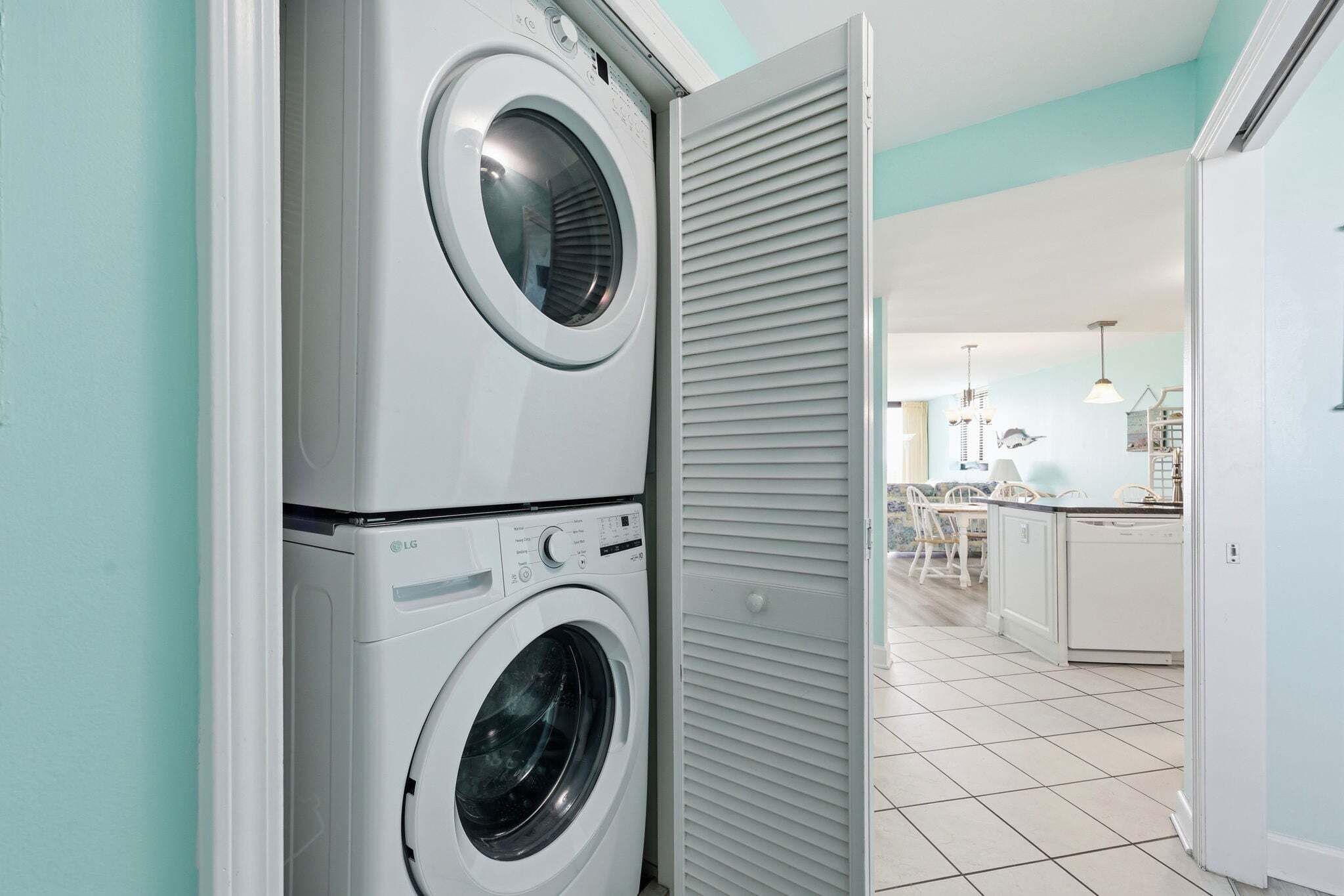 A laundry area with two stacked washing machines, a closed louvered door, and a kitchen area in the background.