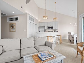 A modern living room with a sofa, a coffee table with magazines, and a kitchen area with white cabinets and a dining table.