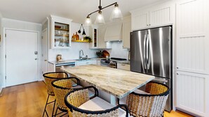 A modern kitchen with a central island, stainless steel appliances, and wicker chairs.
