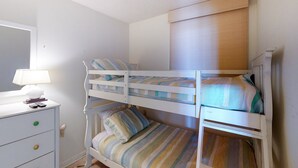 Ett rum med en våningssäng, en vit byrå, en lampa och ett fönster med persienner.