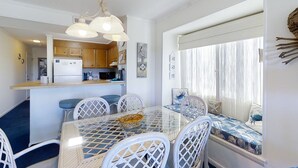 Een eethoek met een glazen tafel en witte stoelen, een keuken op de achtergrond en een raam met gordijnen.