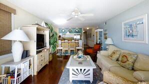 A living room with a TV, white furniture, a ceiling fan, and a dining area with chairs and a table.