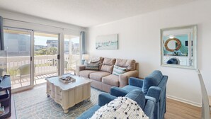 A living room with a beige sofa, a blue armchair, and a coffee table. There is a large window with a view of buildings and a balcony.