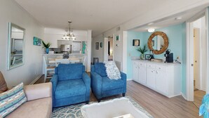 A living room with two blue armchairs, a beige sofa, and a white coffee table. A kitchen is visible in the background with white cabinets and a blue wall.
