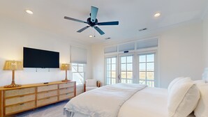 A bedroom with a bed, a ceiling fan, a TV, a wooden console, and large windows. - Soundside Serenity: Perfect Beach Getaway with Water Views, Shared Pool (Atlantic Beach)