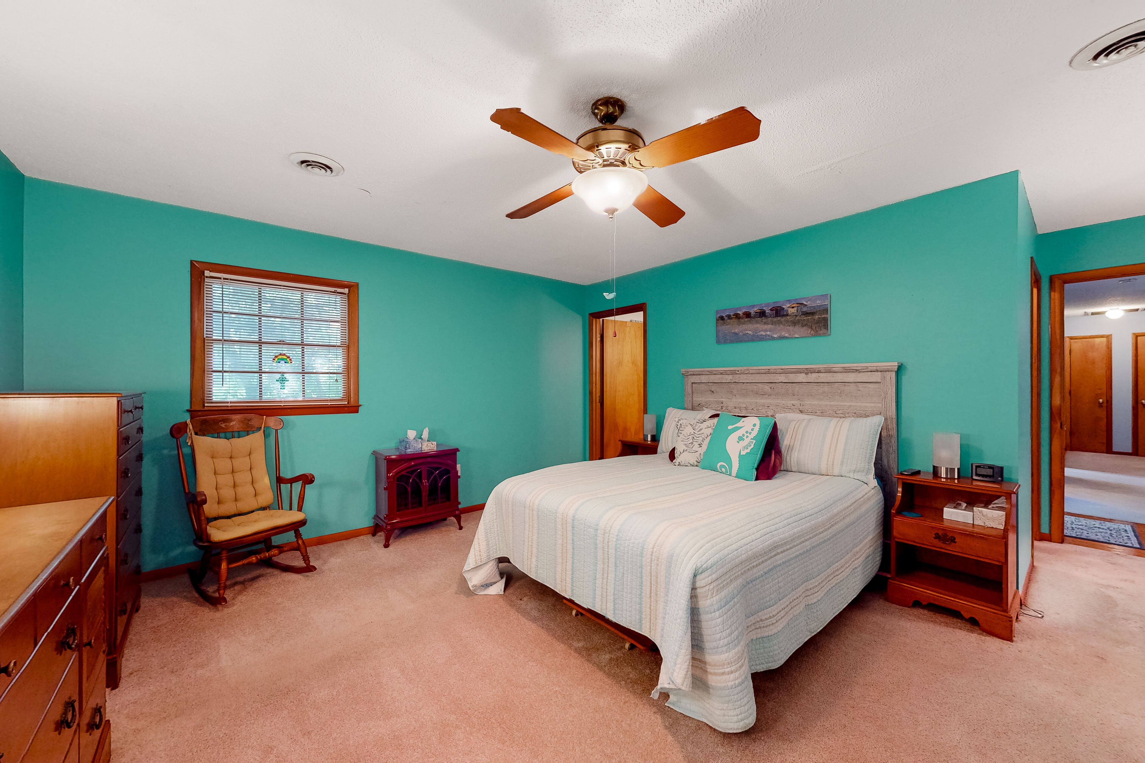 A bedroom with teal walls, a wooden bed with a white comforter, a rocking chair, a wooden nightstand, and a ceiling fan.
