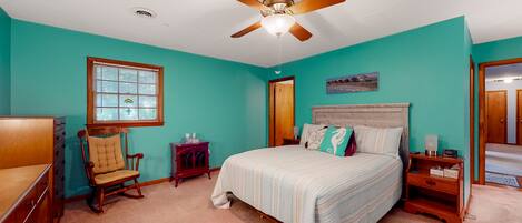 A bedroom with teal walls, a wooden bed with a white comforter, a rocking chair, a wooden nightstand, and a ceiling fan.