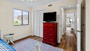 A bedroom with a bed, a TV on a dresser, a window, and a hallway leading to a bathroom. - Cheery coastal condo with balcony, AC, W/D, 2 pools, grills, & beach access (Atlantic Beach)
