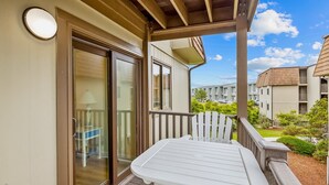 A balcony with a white table and chairs, a sliding glass door, and a view of neighboring buildings. - Cheery coastal condo with balcony, AC, W/D, 2 pools, grills, & beach access (Atlantic Beach)